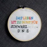 Ein Stickrahmen mit dem Spruch "Das Leben ist zu bunt für schwarz-weiß-denken", kunstvoll mit bunten **Buchstaben sticken** verziert.