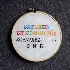 Ein Stickrahmen mit dem Spruch "Das Leben ist zu bunt für schwarz-weiß-denken", kunstvoll mit bunten **Buchstaben sticken** verziert.