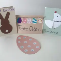 Osterkarten basteln einfach: Hase, Küken und bunte Eier. Ein rosa Ei mit Buchstaben für "Frohe Ostern" liegt vorne.
