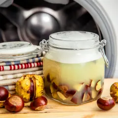 Selbstgemachtes Waschmittel aus Kastanien in einem Glas, neben frischen Kastanien und Wäsche vor einer Waschmaschine.