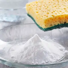 Ein Berg weißes Pulver, das ist Soda, liegt in einer Glasschale. Daneben ein Schwamm und ein Glas Wasser.