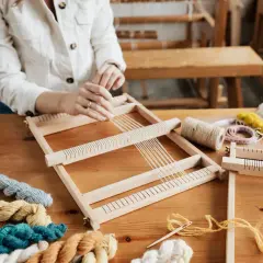 Hände weben auf einem Holzrahmen. Bunte Wollknäuel und Werkzeuge liegen bereit. Tolle Ideen für Erwachsene zum Weben.