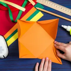 Hände falten orangefarbenes Papier für ein **Gutschein basteln**. Bastelmaterialien wie Bänder, Schere, Kleber und Lineal liegen bereit.