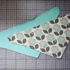 Zwei Stoffteile liegen auf einer Schneidematte. Ein Teil ist mintgrün, das andere hat ein Blumenmuster. Ein Schnittmuster für ein Halstuch Baby.
