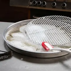Grillrost mit eingebrannten Resten wird in Seifenlauge eingeweicht. Eine Bürste liegt daneben, bereit, den eingebrannten Grillrost zu reinigen.