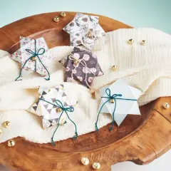 Fünf gefaltete Sternschachteln mit weihnachtlichen Mustern, verziert mit Schnur und Glöckchen, liegen auf einem cremefarbenen Tuch in einer Holzschale.