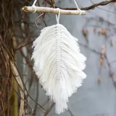 Ein zartes makramee blatt aus weißer Baumwolle hängt an einem Ast. Es wirkt wie ein gefallenes Blatt, das vom Wind getragen wird.