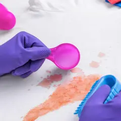 Lila Handschuhe entfernen mit Bürste und Löffel rote Flecken, vielleicht Cranberry-Flecken, von weißem Stoff.