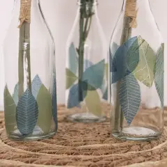 Schöne Serviettentechnik Ideen: Glasflaschen mit Blattmotiven verziert, auf einem geflochtenen Untersetzer.