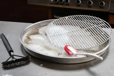 Grillrost mit eingebrannten Resten wird in Seifenlauge eingeweicht. Eine Bürste liegt daneben, bereit, den eingebrannten Grillrost zu reinigen.
