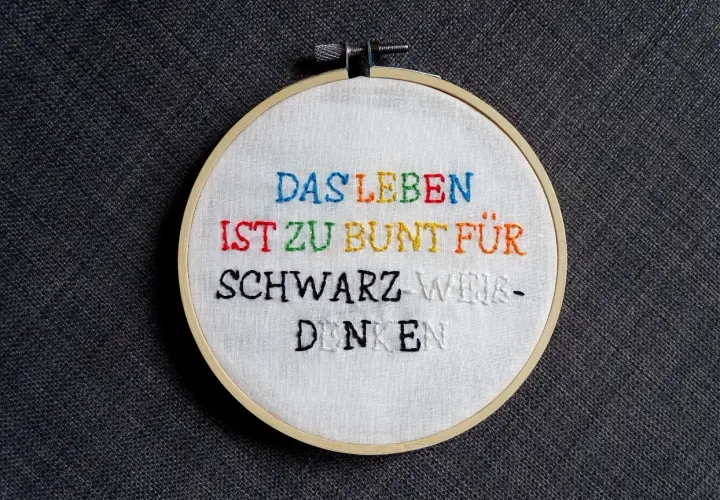 Ein Stickrahmen mit dem Spruch "Das Leben ist zu bunt für schwarz-weiß-denken", kunstvoll mit bunten **Buchstaben sticken** verziert.