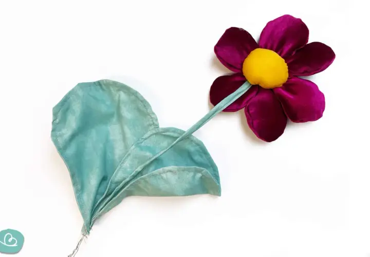 Stoffblumen selber machen: Eine lila Blüte mit gelbem Zentrum und ein grünes Blatt aus Stoff, auf weißem Grund.