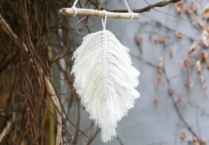 Ein zartes makramee blatt aus weißer Baumwolle hängt an einem Ast. Es wirkt wie ein gefallenes Blatt, das vom Wind getragen wird.