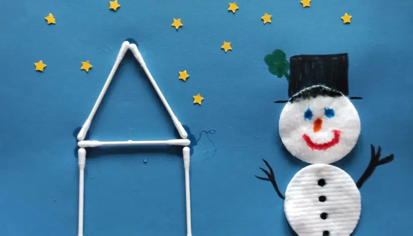 Ein fröhlicher schneemann aus wattepads steht vor einem Haus aus Wattestäbchen. Kleine gelbe Sterne funkeln am blauen Himmel.