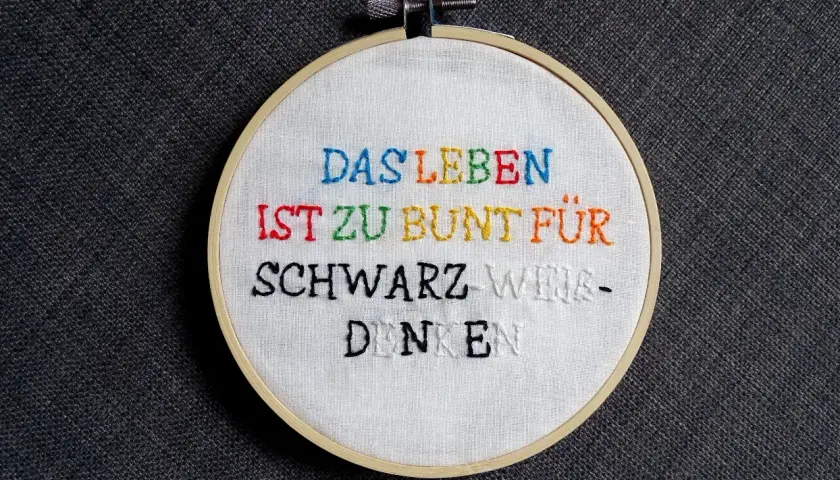 Ein Stickrahmen mit dem Spruch "Das Leben ist zu bunt für schwarz-weiß-denken", kunstvoll mit bunten **Buchstaben sticken** verziert.
