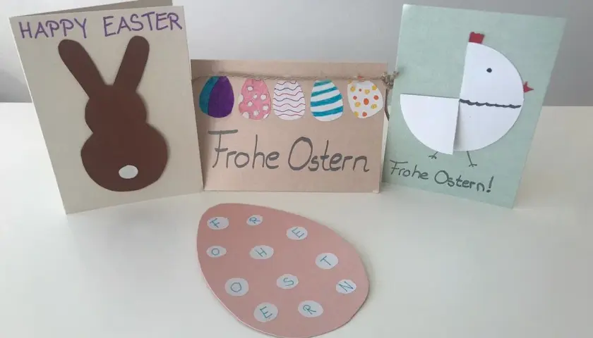 Osterkarten basteln einfach: Hase, Küken und bunte Eier. Ein rosa Ei mit Buchstaben für "Frohe Ostern" liegt vorne.