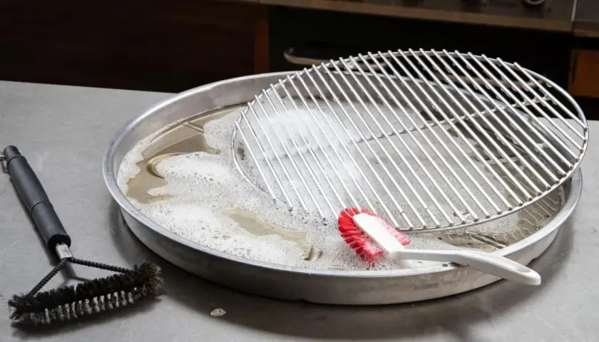 Grillrost mit eingebrannten Resten wird in Seifenlauge eingeweicht. Eine Bürste liegt daneben, bereit, den eingebrannten Grillrost zu reinigen.