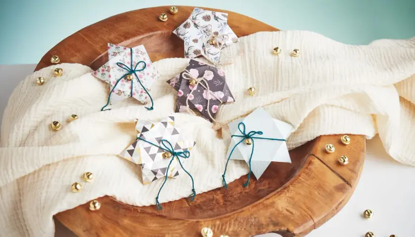 Fünf gefaltete Sternschachteln mit weihnachtlichen Mustern, verziert mit Schnur und Glöckchen, liegen auf einem cremefarbenen Tuch in einer Holzschale.