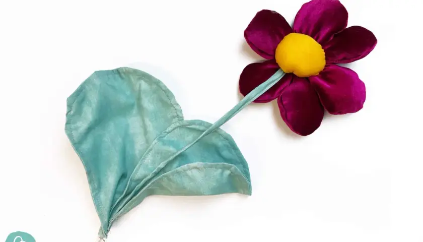 Stoffblumen selber machen: Eine lila Blüte mit gelbem Zentrum und ein grünes Blatt aus Stoff, auf weißem Grund.