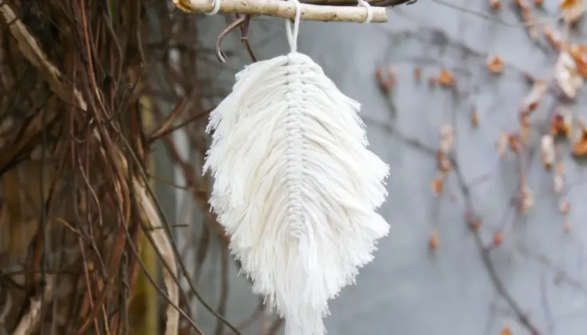 Ein zartes makramee blatt aus weißer Baumwolle hängt an einem Ast. Es wirkt wie ein gefallenes Blatt, das vom Wind getragen wird.