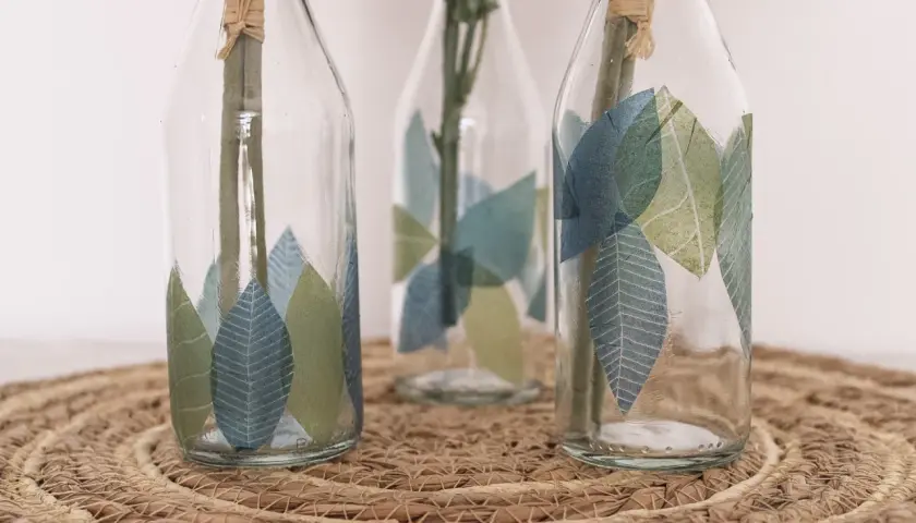Schöne Serviettentechnik Ideen: Glasflaschen mit Blattmotiven verziert, auf einem geflochtenen Untersetzer.