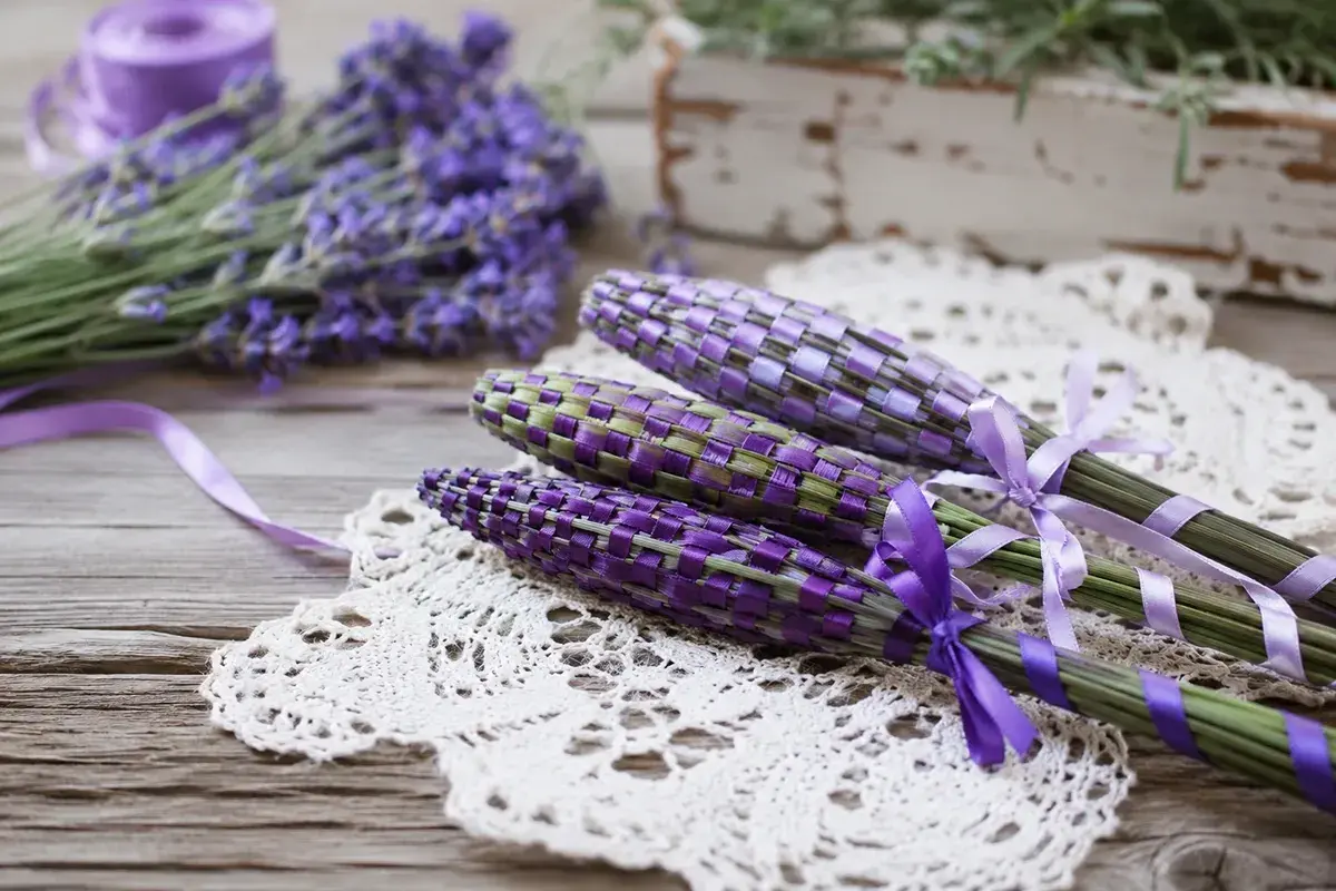 Drei geflochtene Lavendel-Stäbe mit lila Bändern auf einer Spitzendecke. Im Hintergrund ein Bund frischer Lavendel.