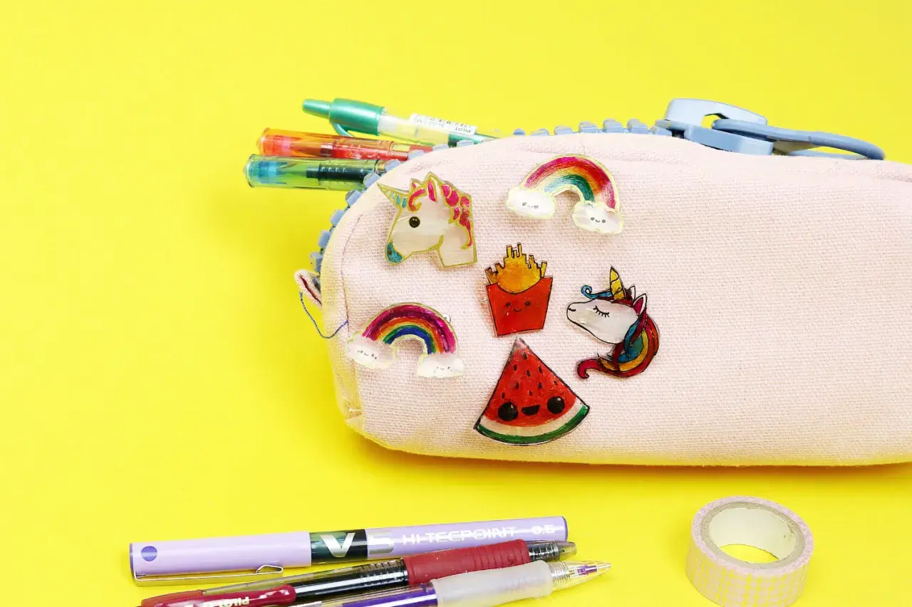 Bunte Schrumpffolie Ideen: Einhorn, Regenbogen, Pommes & Wassermelone zieren eine Federtasche. Stifte & Klebeband liegen daneben.