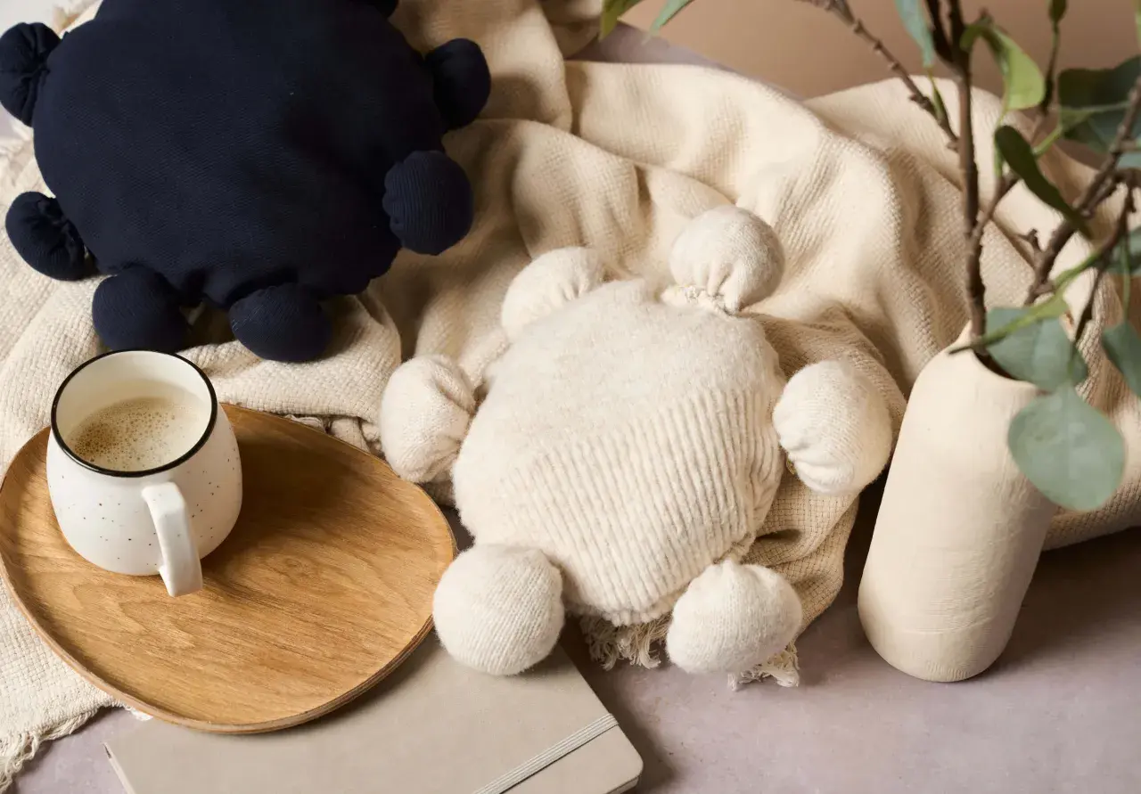 Zwei Kuscheltiere, eines dunkelblau, das andere cremefarben, liegen auf einer Decke. Ein Becher Kaffee steht auf einem Holztablett. Kreatives **Pullover Upcycling** für süße Spielzeuge.