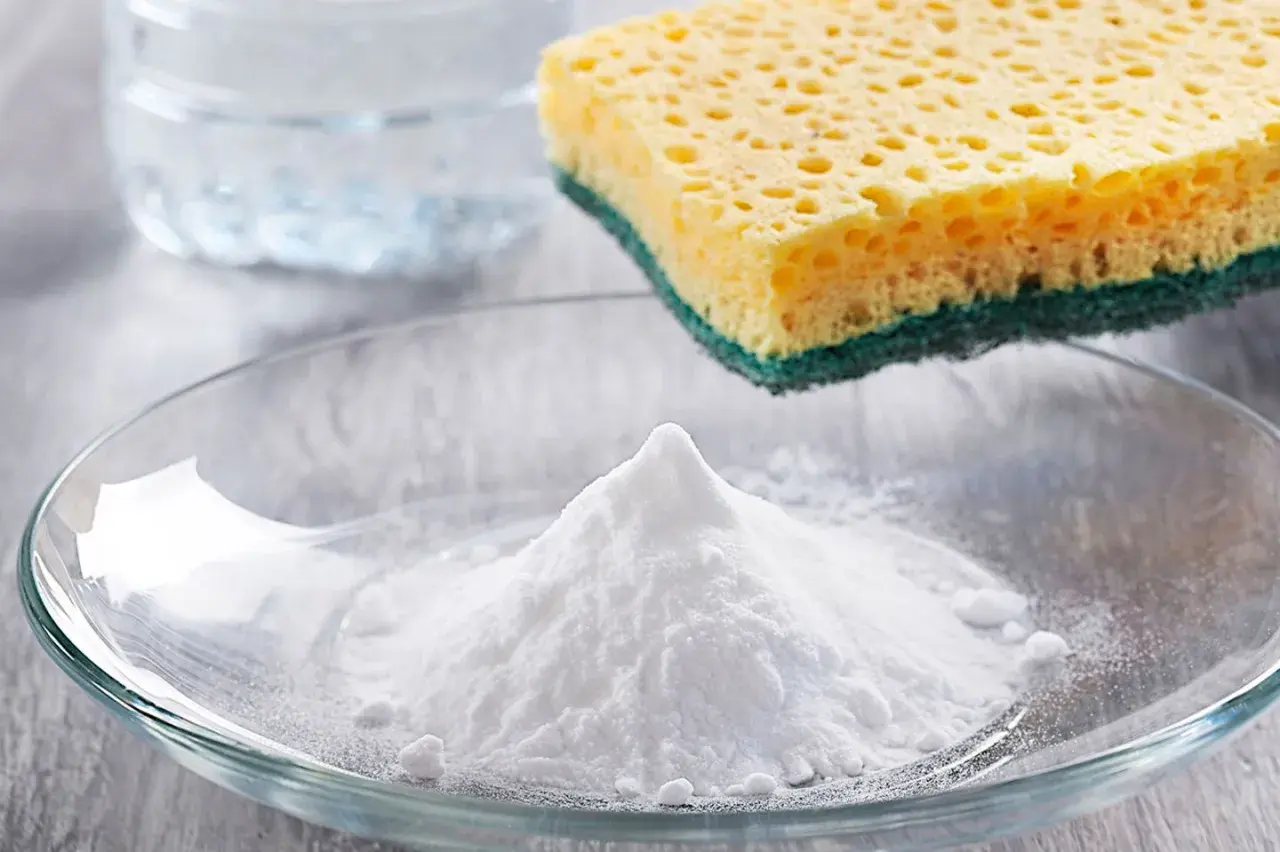 Ein Berg weißes Pulver, das ist Soda, liegt in einer Glasschale. Daneben ein Schwamm und ein Glas Wasser.