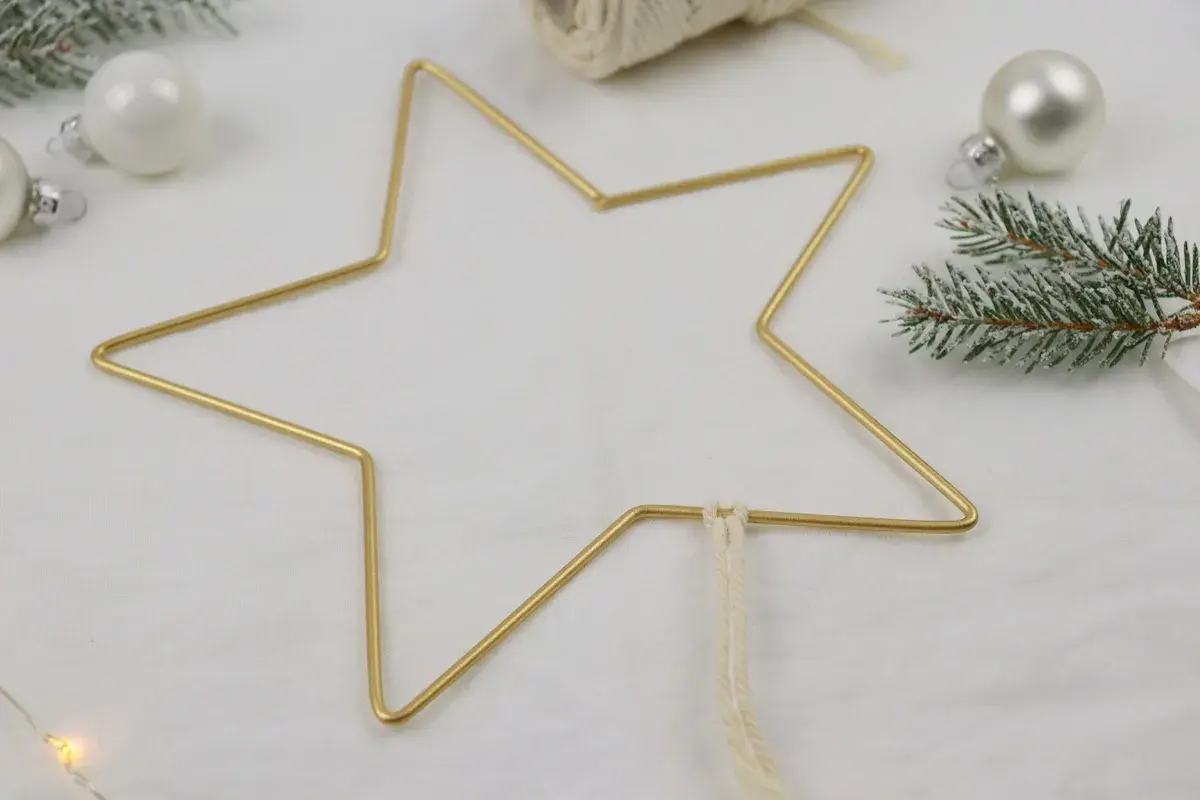 Goldener Sternrahmen für Makramee Weihnachten, umgeben von Tannenzweigen und weißen Kugeln.