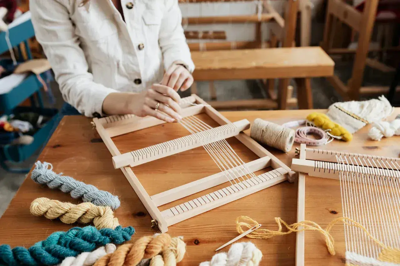 Hände weben auf einem Holzrahmen. Bunte Wollknäuel und Werkzeuge liegen bereit. Tolle Ideen für Erwachsene zum Weben.