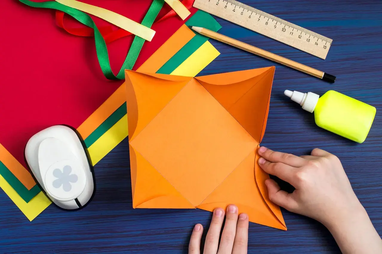 Hände falten orangefarbenes Papier für ein **Gutschein basteln**. Bastelmaterialien wie Bänder, Schere, Kleber und Lineal liegen bereit.