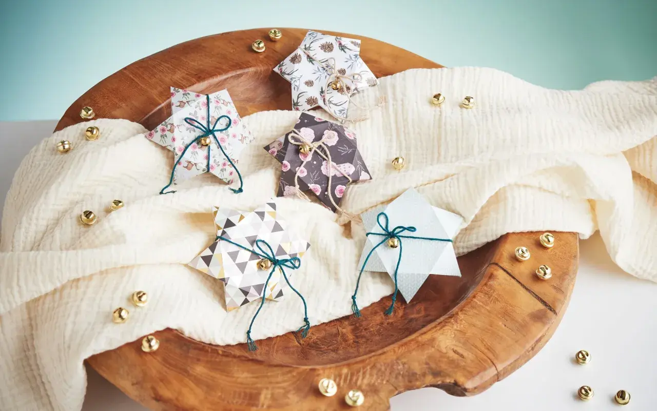 Fünf gefaltete Sternschachteln mit weihnachtlichen Mustern, verziert mit Schnur und Glöckchen, liegen auf einem cremefarbenen Tuch in einer Holzschale.