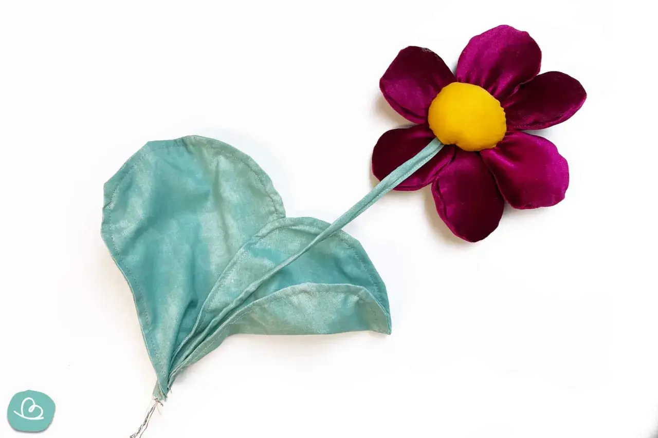 Stoffblumen selber machen: Eine lila Blüte mit gelbem Zentrum und ein grünes Blatt aus Stoff, auf weißem Grund.