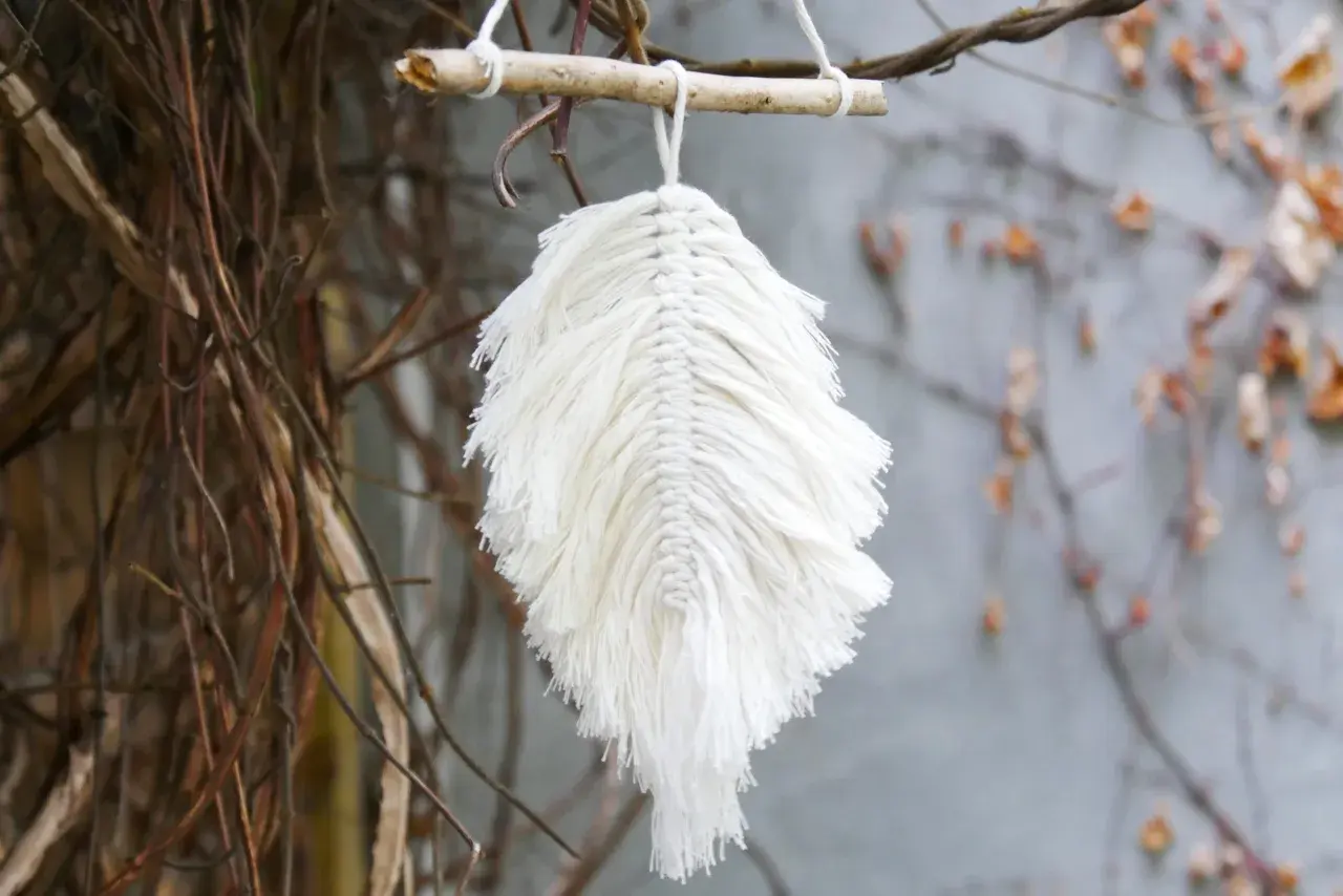 Ein zartes makramee blatt aus weißer Baumwolle hängt an einem Ast. Es wirkt wie ein gefallenes Blatt, das vom Wind getragen wird.