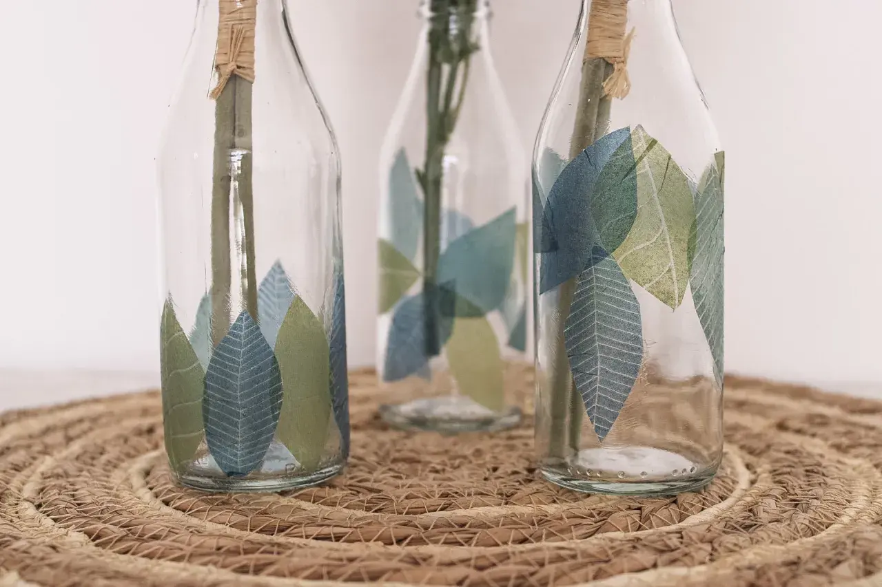 Schöne Serviettentechnik Ideen: Glasflaschen mit Blattmotiven verziert, auf einem geflochtenen Untersetzer.