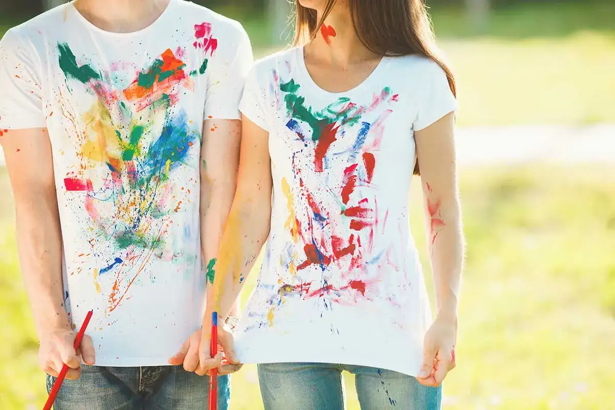 Bunte Farbspritzer auf weißen T-Shirts. Ein Paar hält Pinsel, bereit, die Textilfarbe zu entfernen und neue Kunst zu schaffen.
