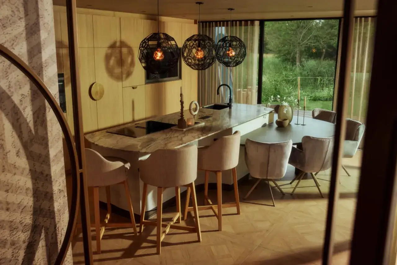 Moderne Essecke mit K&uuml;cheninsel, Barhockern und Esstisch vor bodentiefen Fenstern. Holzm&ouml;bel und ged&auml;mpfte Farben schaffen eine warme Atmosph&auml;re.