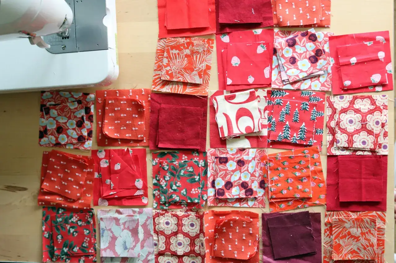 Viele rote Stoffquadrate mit Mustern wie Blumen, Erdbeeren und Bäumen liegen bereit für neue patchwork ideen. Eine Nähmaschine ist im Hintergrund zu sehen.