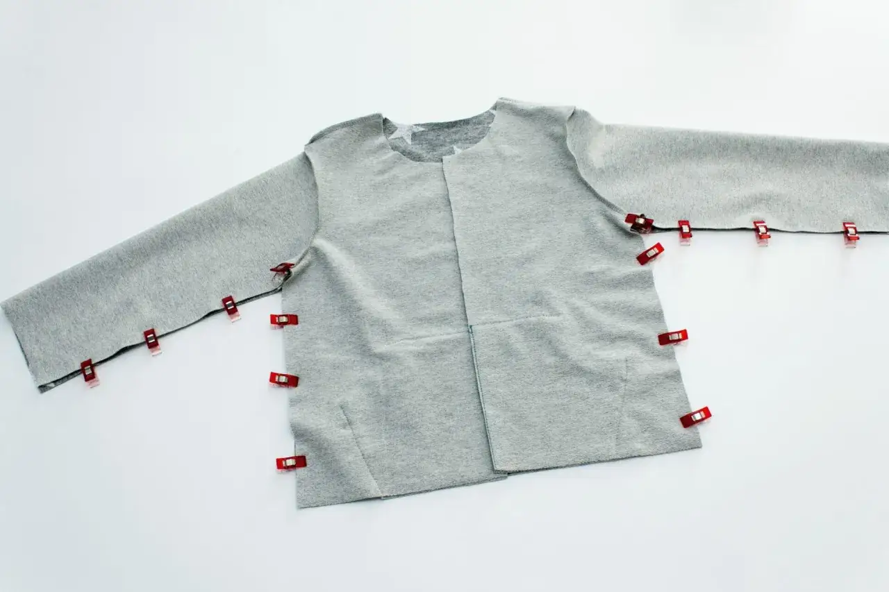 Graues Stoffteil einer Kinderjacke mit roten Klammern, bereit f&uuml;r ein kostenloses Schnittmuster einer Kapuzenjacke.