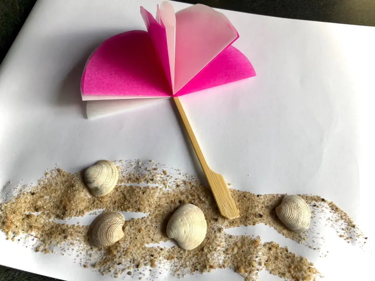 Ein selbstgebastelter Sonnenschirm aus Papier und Holzspieß steht auf Sand mit Muscheln. Perfekt für eine Bastelidee zum Thema Sommer.
