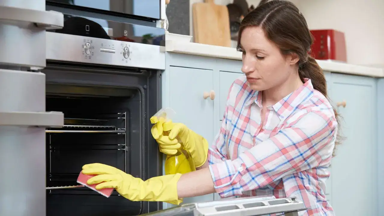 Frau mit gelben Handschuhen reinigt Backofen mit Sprühflasche und Schwamm. Sie macht Backofenreiniger selber.