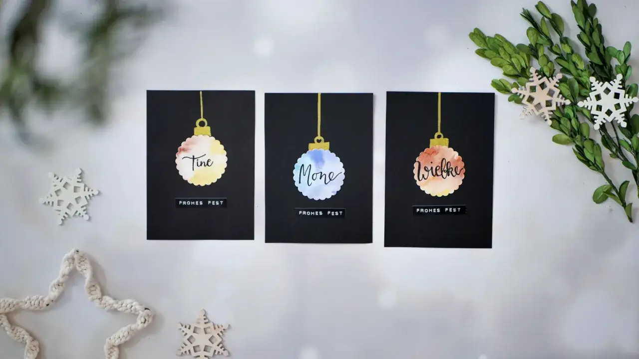 Moderne weihnachtskarten basteln: drei schwarze Karten mit Aquarell-Christbaumkugeln und Namen. Dekoriert mit Schneeflocken und Grün.