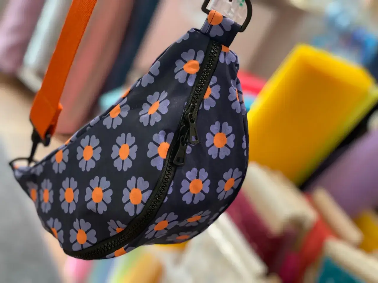 Blumige Bauchtasche mit orangefarbenem Gurt. Das Schnittmuster zeigt lila Blüten mit orangefarbenen Zentren auf dunkelblauem Grund.