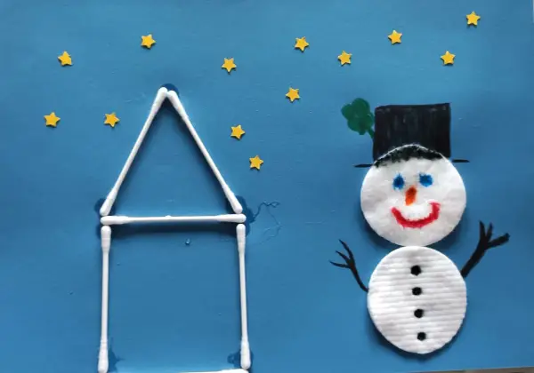 Ein fröhlicher schneemann aus wattepads steht vor einem Haus aus Wattestäbchen. Kleine gelbe Sterne funkeln am blauen Himmel.